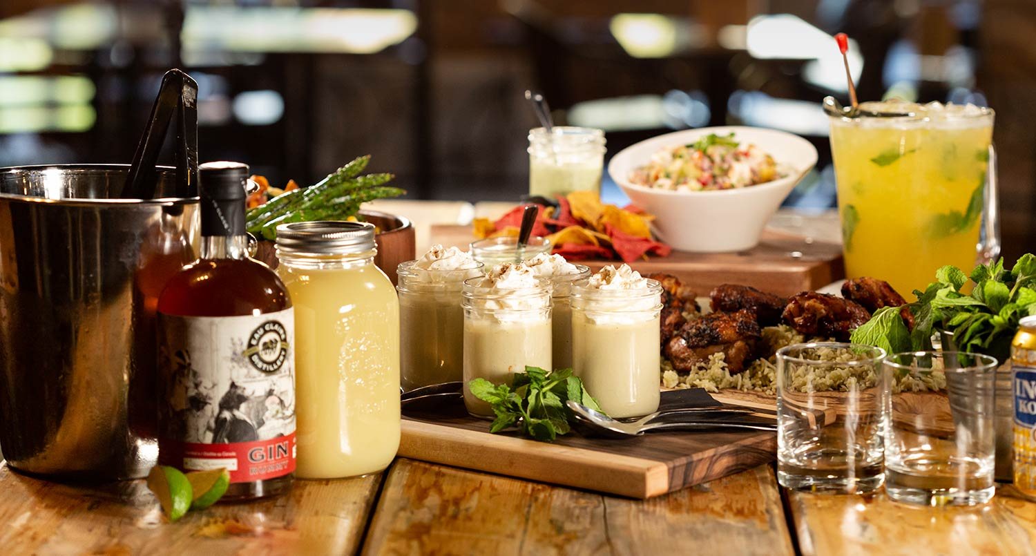 A Bo's spread on a wood table: jars of dessert, wings over rice, a pitcher with mint, gin and a salad